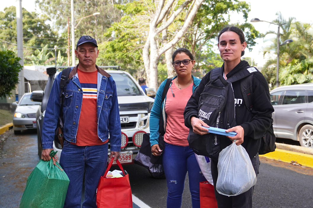 La imagen muestra a tres personas de pie en una calle, cargando bolsas y mochilas. Se trata de un …